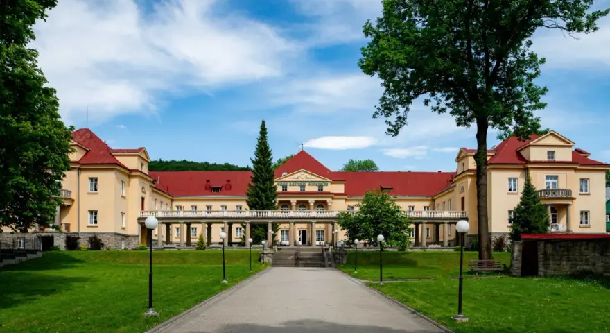 Eleganckie, żółte budynki z czerwonymi dachami, przypominające stare łazienki Krynica Zdrój, otoczone zielonym ogrodem.