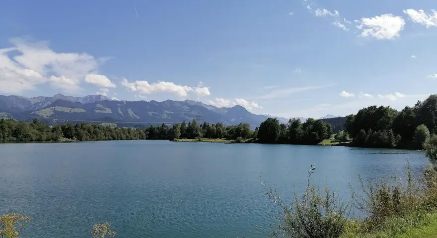Blauer Himmel über dem Sonthofener See, umgeben von grünen Bäumen und majestätischen Bergen.