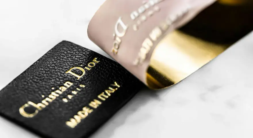 Etichetta in pelle nera con logo Christian Dior Paris e "Made in Italy" dorato, accanto a un nastro Dior francese con finitura oro lucido.