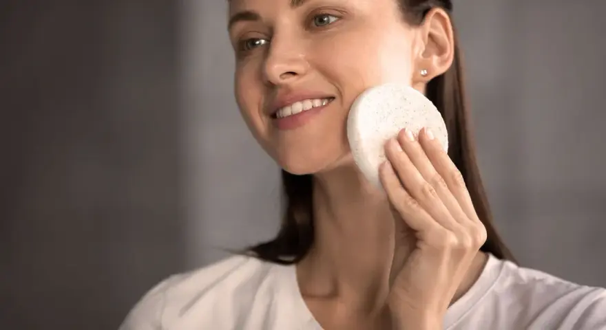 Une femme souriante utilise une éponge naturelle pour se laver le visage. Une méthode douce pour un nettoyage en douceur.