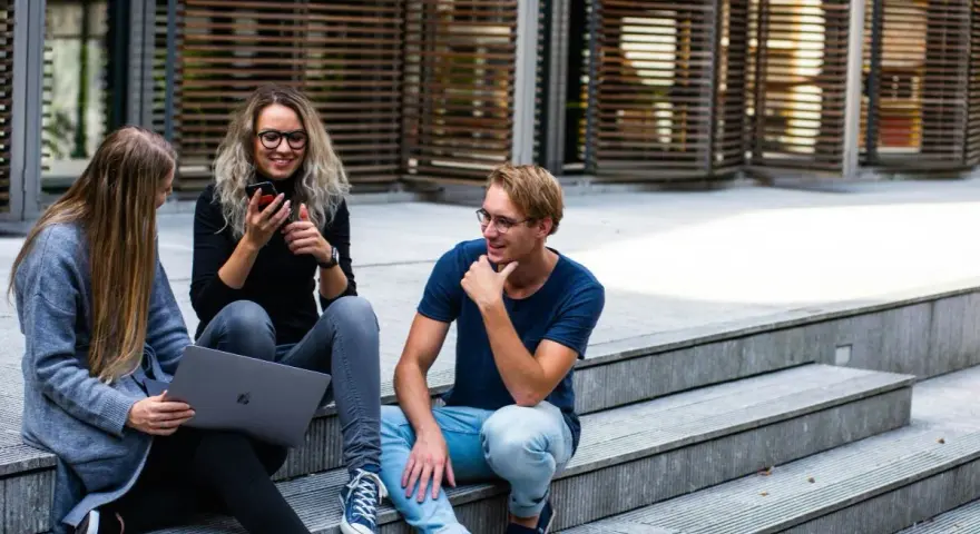 Trójka studentów, w tym jedna osoba na studiach lingwistycznych, siedzi na schodach, korzystając z laptopa i telefonu.