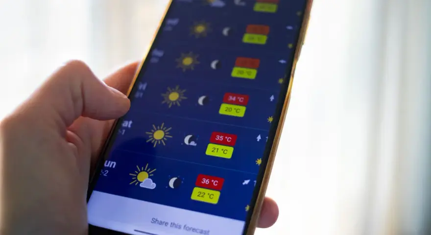 Dłoń trzyma smartfon z otwartą aplikacją pogoda, pokazującą prognozę z wysokimi temperaturami.