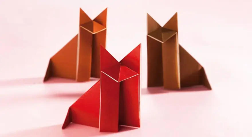 Drei origami Füchse, einer rot, zwei braun, sitzen auf rosa Untergrund.