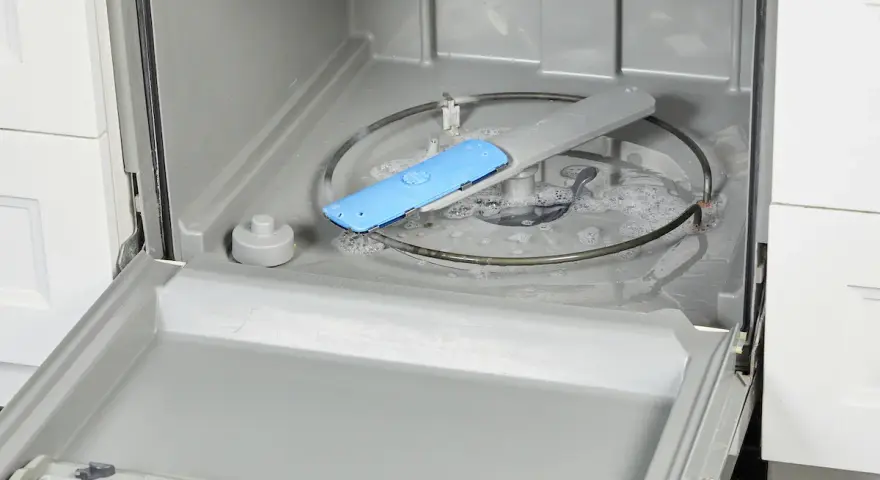 Water and suds fill the bottom of a dishwasher, with a blue spray arm component visible. This image shows a step in how to fix a clogged dishwasher.