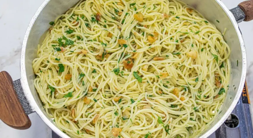 Espaguetis al ajillo recién hechos, con trocitos de ajo dorado y perejil fresco, servidos en una sartén metálica.