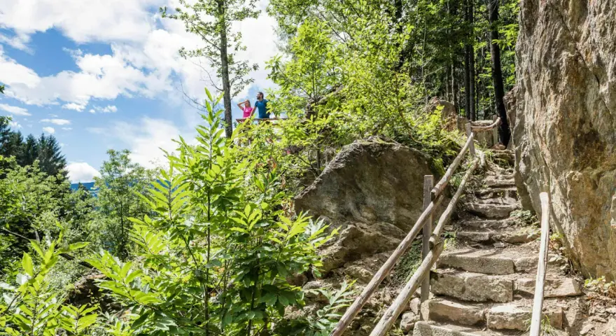 Ein Paar genießt die Aussicht vom Schenner Waalweg. Steintreppen führen durch üppige Vegetation und Felsen.