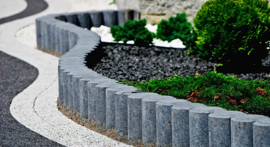 Eleganckie obrzeża trawnikowe z kostki betonowej tworzą falistą linię, oddzielając zadbane rabaty z iglakami od ścieżki.