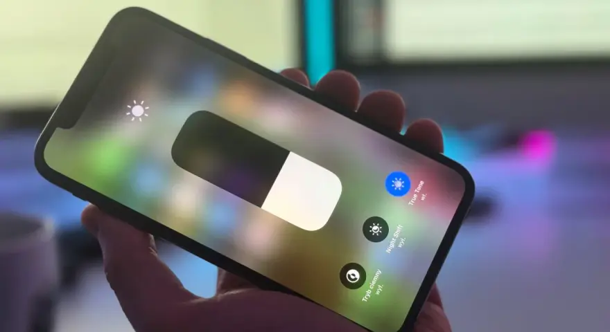 Dłoń trzyma iPhone'a z włączonym panelem sterowania. Widoczny suwak jasności i ikona True Tone.