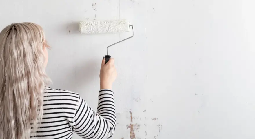Mujer con cabello rubio y camiseta a rayas está a punto de empapelar pared con rodillo blanco.