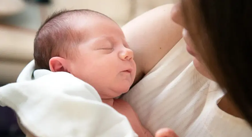 Śpiące niemowlę w ramionach mamy. Warto sprawdzić, ile powinno jeść niemowlę -- tabela, by zapewnić mu najlepszy start.