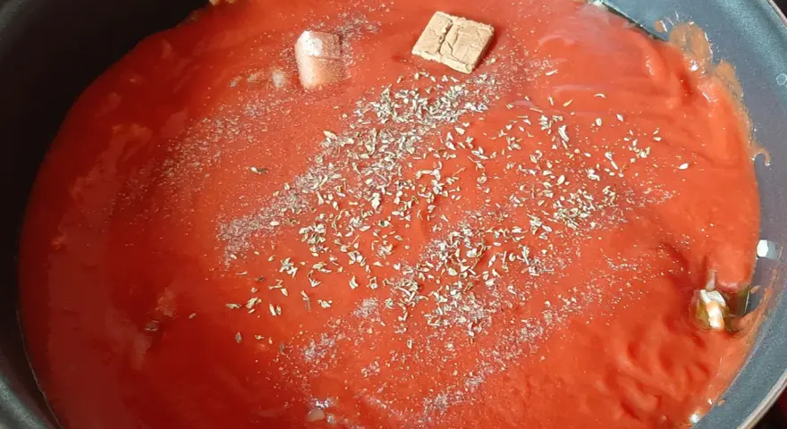 Un coulis de tomates mijote dans une poêle, agrémenté d'herbes et d'un cube de bouillon.