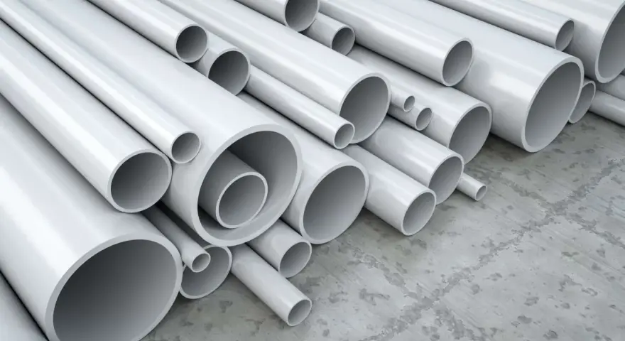 A pile of white pipes, many of which are made of vinyl, a type of plastic, are stacked on a concrete floor.