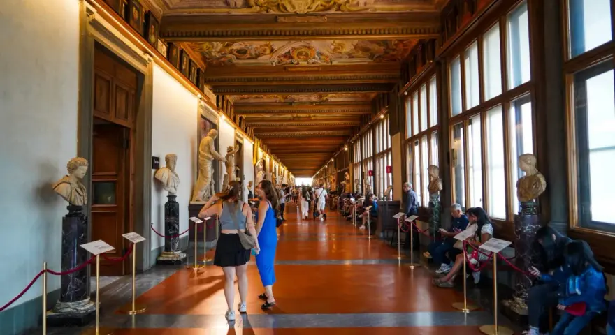 Wnętrze muzeum Uffizi z rzeźbami i malowanym sufitem. Ludzie spacerują i odpoczywają wzdłuż korytarza.