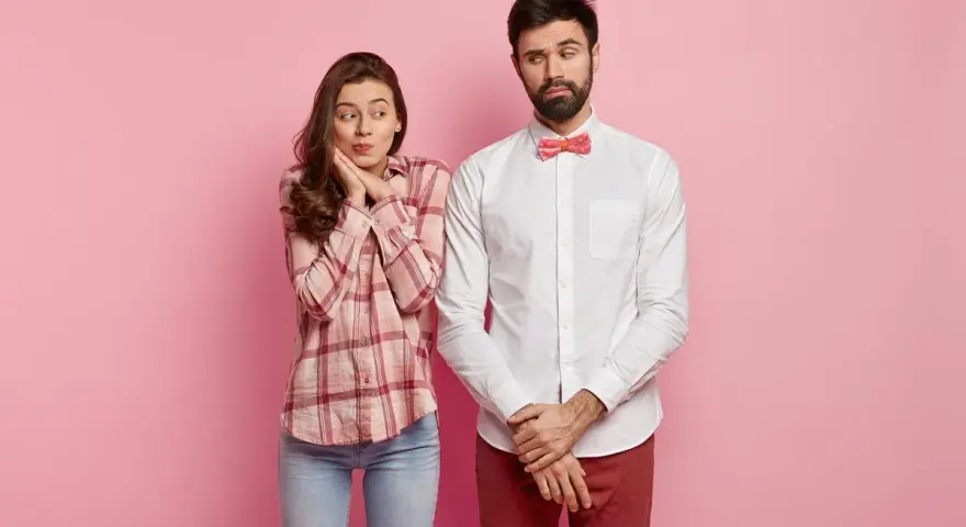 Coppia su sfondo rosa. Lei, con camicia a quadri e jeans, sembra timida. Lui, con camicia bianca e papillon, è pensieroso. Come vestirsi al primo appuntamento uomo?