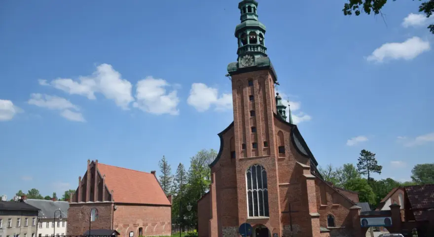 Majestatyczna kolegiata w Kartuzach z cegły, z zieloną wieżą i zegarem, na tle błękitnego nieba z chmurami.