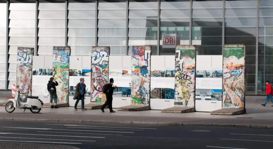 Bunte Reste des Berliner Mauer-Denkmals vor dem Bahnhof Potsdamer Platz. Menschen spazieren vorbei, ein Roller parkt daneben.
