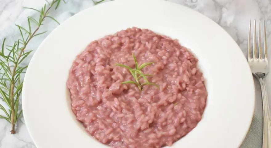 Różowe risotto z prawdziwkami i rozmarynem na białym talerzu, obok widelca i kieliszka wina.
