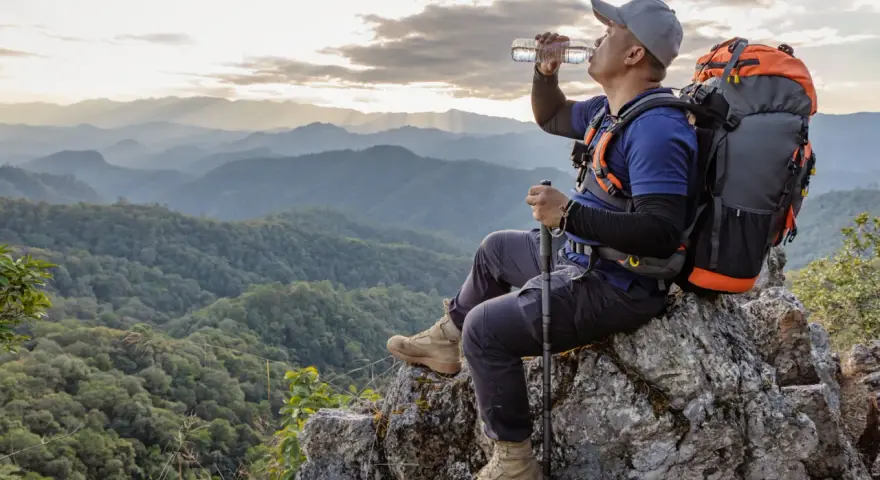 Backpacker drinks water on a rocky outcrop, contemplating how much water to bring backpacking for this stunning mountain vista.