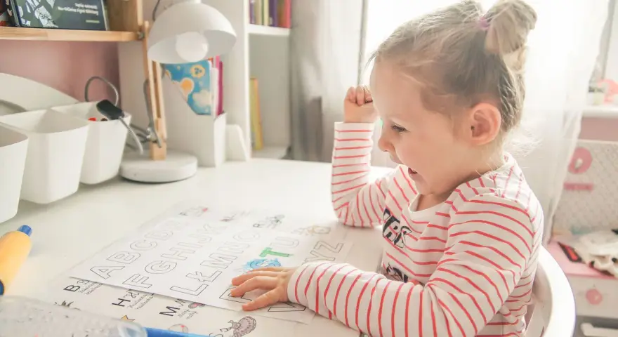 Mała dziewczynka z zapałem uczy się literek dla 3 latka. Na biurku ma kolorowanki z alfabetem i flamastry.