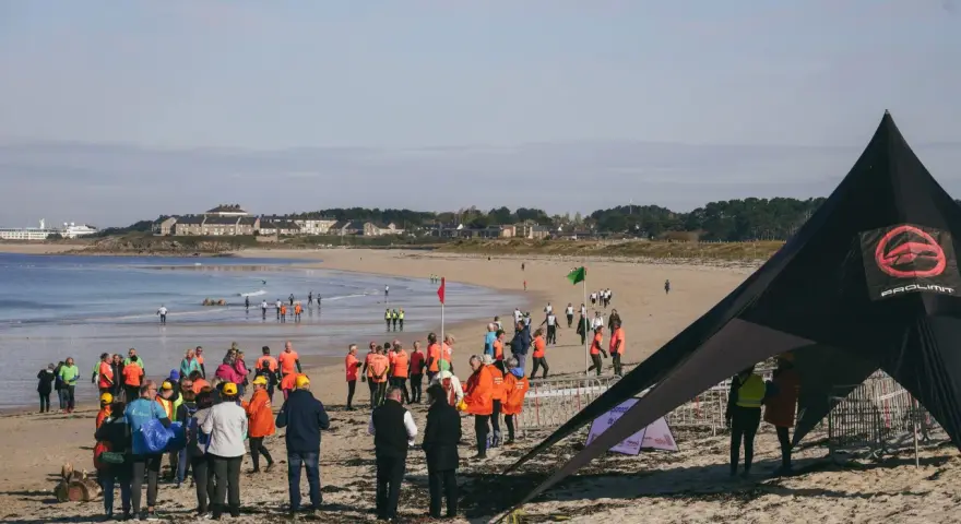 Rassemblés sur la plage pour une course de longe côte, des participants en tenues colorées attendent le départ sous un ciel clair.