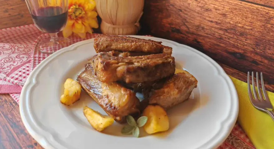 Succulente costine di maiale in padella, dorate e saporite, servite con patate arrosto e un rametto di salvia.