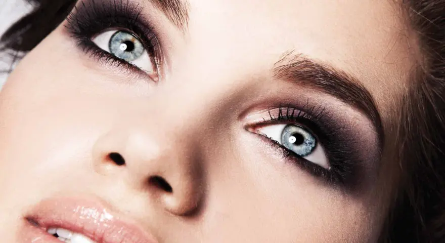 Occhi azzurri intensificati da un trucco smokey eyes audace, con labbra lucide e un accenno di sorriso.