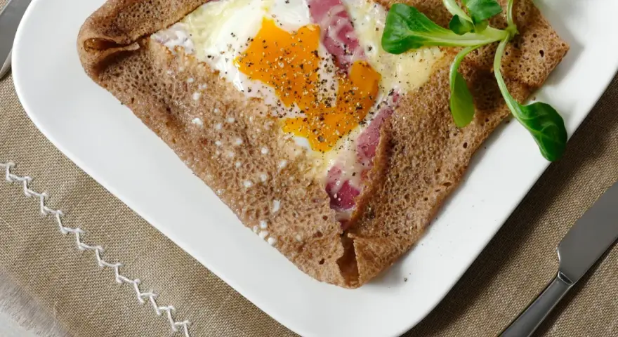Une galette bretonne garnie d'un œuf, de jambon et de fromage, poivrée et décorée d'une feuille verte. Un délice de french crepes !