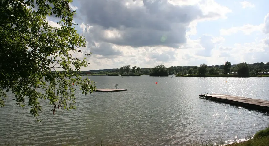 Ein sonniger Tag am Oberrieder Weiher Camping. Ein Steg mit Leiter lädt zum Baden ein, während im Hintergrund Bäume und eine idyllische Landschaft zu sehen sind.