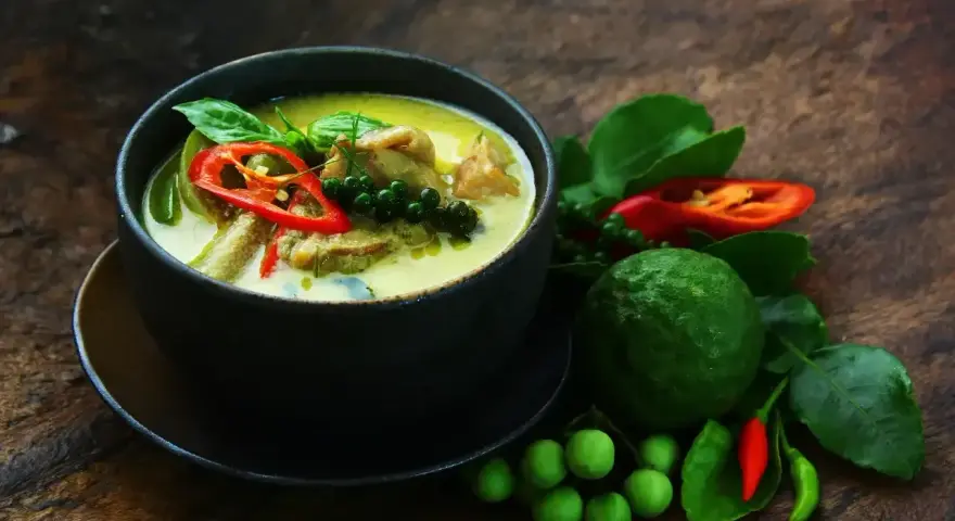 Aromaticzne zielone curry z kawałkami mięsa, papryczką chili, zielonym pieprzem i liśćmi bazylii. Obok miski świeże składniki: limonka kaffir, pomidorki i papryczki chili.