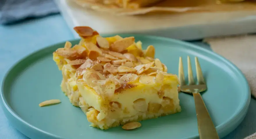 Ein Stück schneller Apfelkuchen mit Schmand und Pudding, bestreut mit Mandelblättchen und Zimt, auf einem türkisfarbenen Teller.