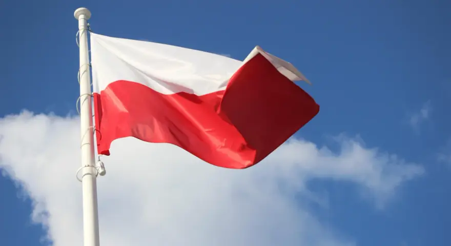 Biało-czerwona bandera polska powiewa na maszcie na tle błękitnego nieba z chmurami.