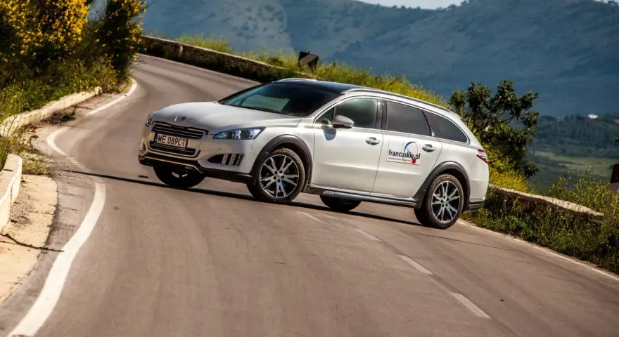 Białe kombi Peugeot 508 RXH na krętej drodze w górach.