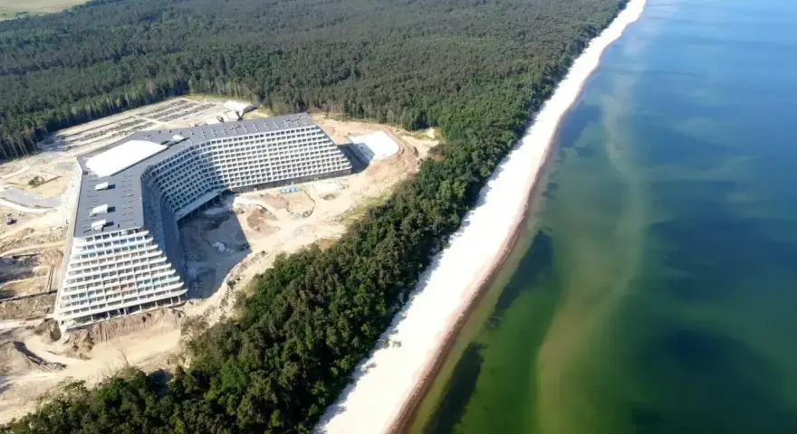 Nowy hotel Gołębiewski w budowie, majestatycznie wznoszący się nad piaszczystą plażą i lasem sosnowym, tuż przy turkusowym morzu.