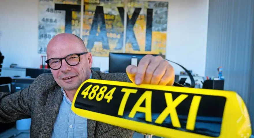 Mann mit Brille hält ein gelbes Taxi-Schild mit der Nummer 4884. Im Hintergrund Kunstwerke mit "TAXI" und der Zahl 1884.