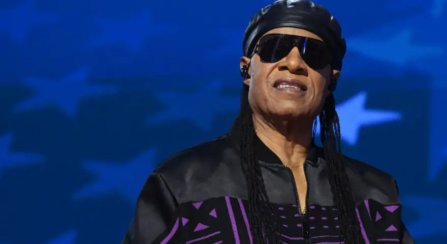 Stevie Wonder heute, mit Sonnenbrille und schwarzer Kappe, trägt eine Jacke mit lila Muster vor blauem Sternenhintergrund.