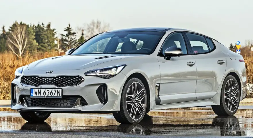 Elegancka Kia Stinger 2025 w kolorze srebrnym, zaparkowana na mokrej nawierzchni, z drzewami w tle.