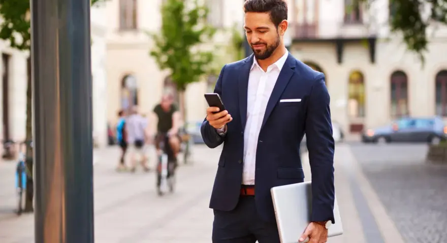 Mężczyzna w eleganckim stroju biznesowym męskim sprawdza telefon, trzymając laptopa.