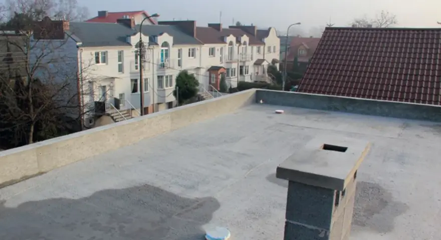 Nowy, betonowy dach płaski z kominem i widokiem na szereg domów z dachówką.