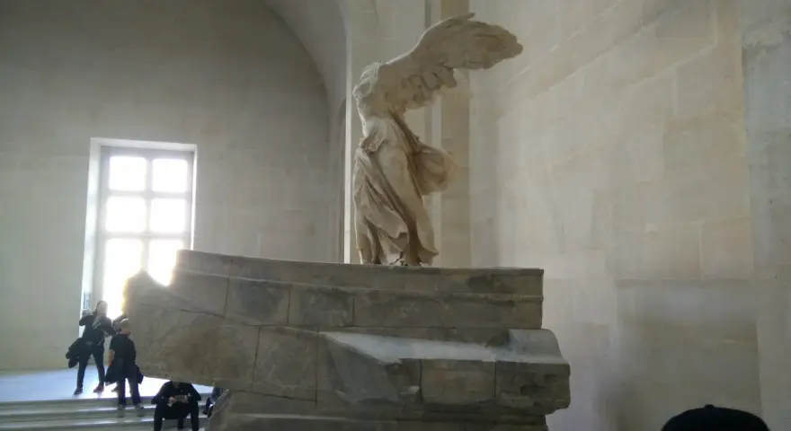 Skrzydlata Nike, symbol zwycięstwa, niczym mit, unosi się nad kamiennym postumentem.