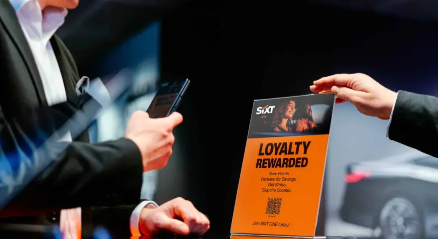 Ein Mann hält ein Smartphone, während eine Hand ein orangefarbenes Schild mit der Aufschrift "Loyalty Rewarded" und dem SIXT ONE Logo hochhält.