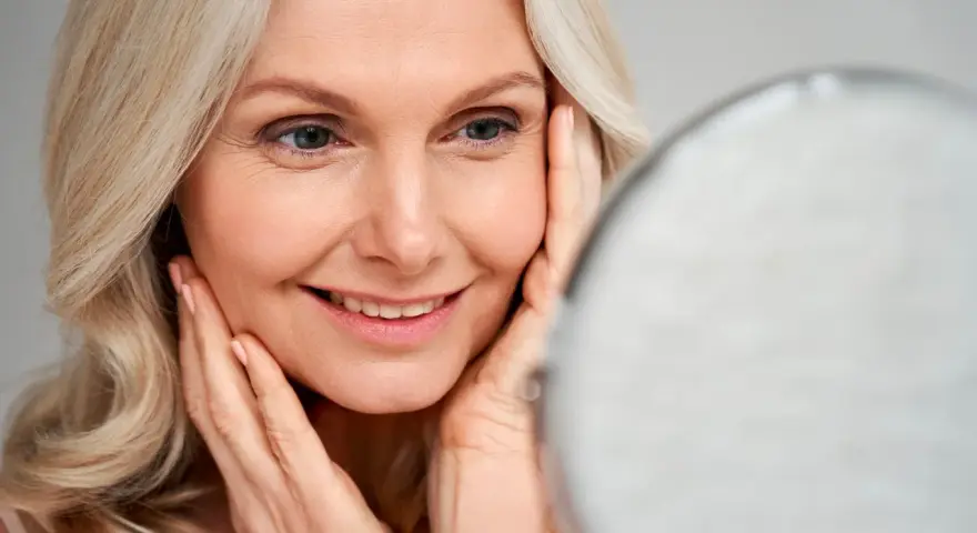Une femme blonde sourit en se regardant dans un miroir, ses mains caressant son visage. Le maquillage paupière tombante avant après est subtil.