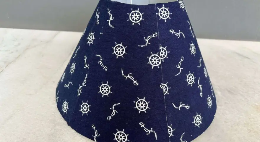 Un abat-jour conique bleu marine orné de motifs d'ancre et de roue de gouvernail. Idéal pour fabriquer un abat-jour en tissu plissé avec une touche marine.