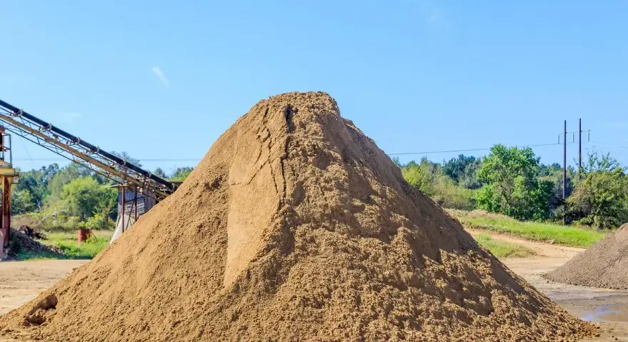 Ogromna hałda piasku na placu budowy, zastanawiasz się, ile kosztuje tona piasku? W tle taśmociąg i drzewa.