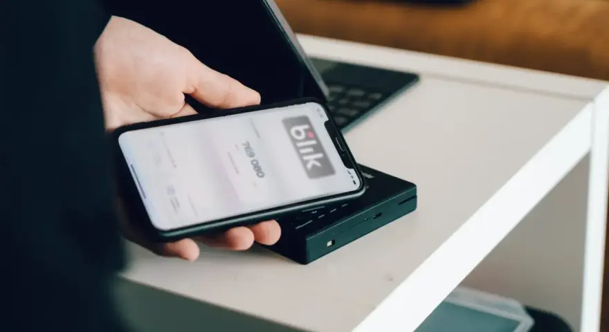 Dłoń trzyma smartfon z aplikacją Blik i kodem transakcji nad terminalem płatniczym.