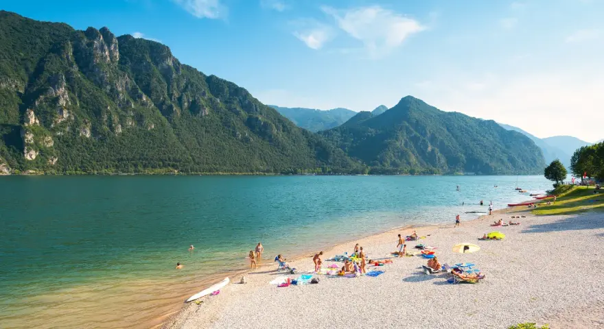 Spiaggia affollata sui laghi in Piemonte, ideale per una gita in moto. Montagne verdi e acqua azzurra.