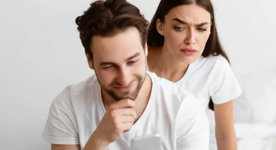 Une femme regarde par-dessus l'épaule de son partenaire qui utilise son téléphone, l'air suspicieuse. Elle essaie de calmer sa jalousie.