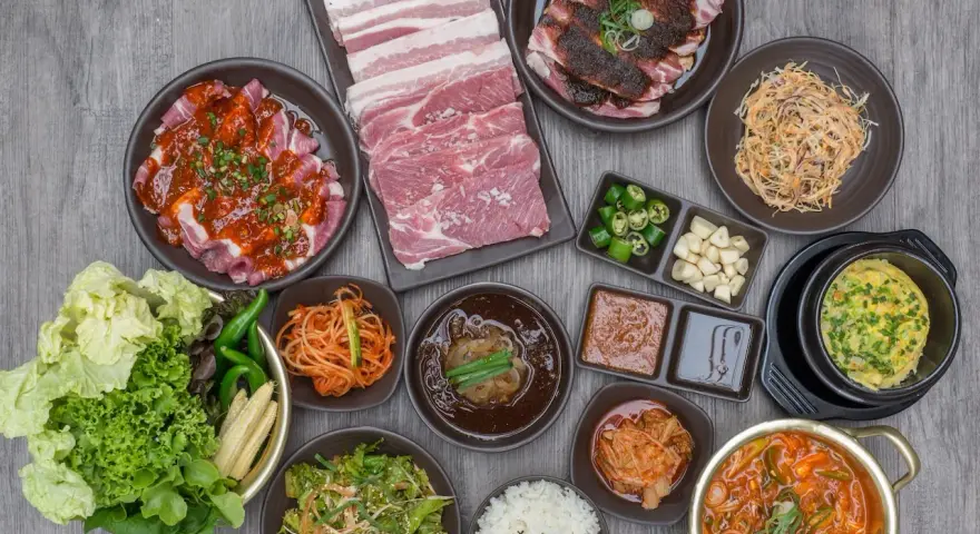 Obfity zestaw do korean bbq: surowe i marynowane mięso, kimchi, sałatki, ryż i dodatki.