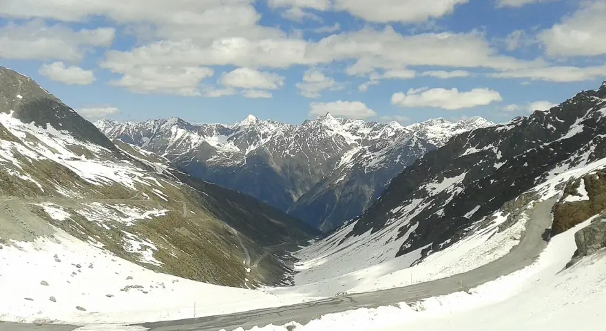 Strada tortuosa sulla Ötztaler Gletscherstraße, tra montagne innevate e cielo azzurro con nuvole bianche.