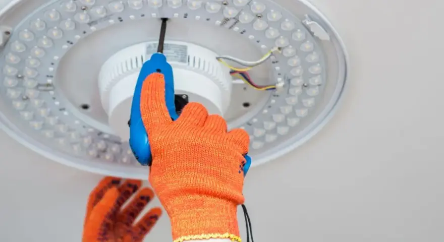 Ręka w pomarańczowej rękawicy odkręca śrubę plafonu LED. Instrukcja jak zdjąć plafon krok po kroku.