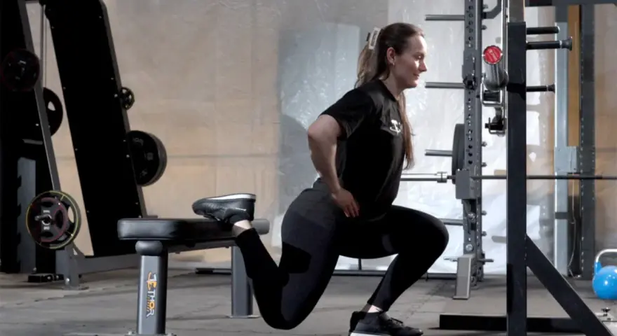 Kvinna utför en split squat i ett gym. Hennes ena fot vilar på en bänk bakom henne.